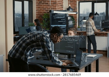 Senior developer codes on a laptop at a desk with multiple screens parsing software, compiling programming language and reviewing algorithm results.