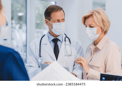 Senior doctor consulting female adult patient near reception desk in hospital hall. Coronavirus concept. Diagnosis, treatment, medications discussion - Powered by Shutterstock