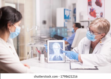 Senior Doctor With Face Mask Against Covid-19 Pointing At Brain Trauma On Table Computer During Patient Examination. Young Woman With Skull Injury.