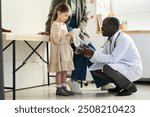 Senior doctor kneeling and assisting young girl holding white stuffed animal in medical office environment with diverse people present and medical equipment surrounding