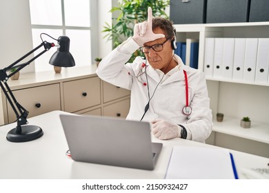 Senior Doctor Man Working On Online Appointment Making Fun Of People With Fingers On Forehead Doing Loser Gesture Mocking And Insulting. 