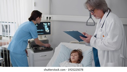 Senior Doctor And Nurse Checking Sick Child Patient Sleeping In Hospital Bed. Medical Team Checking Vital Signs Of Ill Preteen Boy Lying Unconscious In Hospital Ward