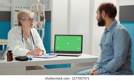 Senior Doctor And Patient Looking At Laptop With Greenscreen At Checkup Visit. Man And Woman Using Isolated Template With Blank Chromakey Background And Mockup Copyspace On Display.