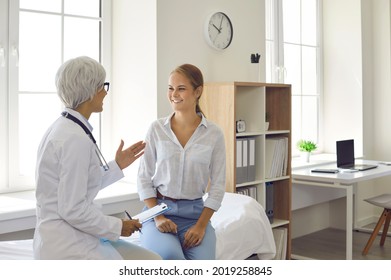 Senior doctor talking to young female patient. Happy cheerful smiley beautiful woman listening to her physician while sitting on examination bed during health consultation in modern clinic or hospital - Powered by Shutterstock