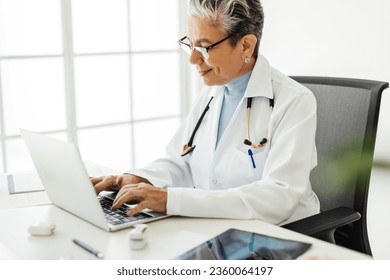 Senior doctor using a laptop to access medical software, which allows her to practice efficiently. Female physician working with technology in her office, wearing a white lab coat and a stethoscope. - Powered by Shutterstock