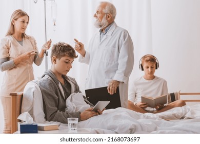 Senior Doctor Wearing Medical Apron And Glasses Talking With Teenage Patients Sitting On Hospital Beds