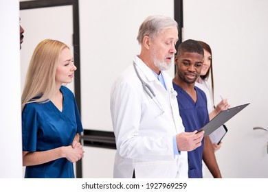 Senior Doctor In White Uniform Consulting Young Doctors Therapists Using Clipboard In Hospital, Multidisciplinary Medical Team In White Coats.