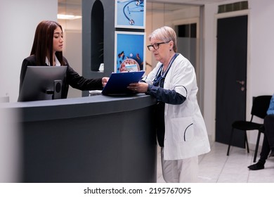 Senior Doctor Working With Asian Receptionist To Plan Checkup Report Papers At Hospital Reception Desk. Medic Taking To Clinic Worker At Counter About Medical Forms And Healthcare Insurance Support.