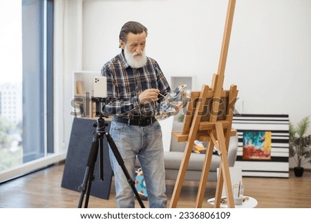 Senior drawing expert making live streaming from spacious apartment. Male artist recording own video tutorial on smartphone about art painting using tripod. Freelancer sharing online favourite job.