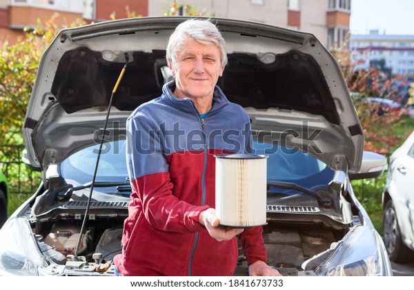 Senior driver holding new\
clean air cartridge for engine airbox. Car with opened hood is on\
background