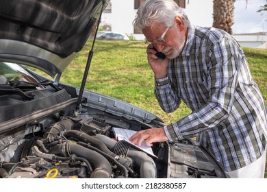 Senior Driver Man Stopped In The Street For A Car Breakdown With The Hood Open Looks At The Instruction Booklet Talking On Phone For Roadside Assistance