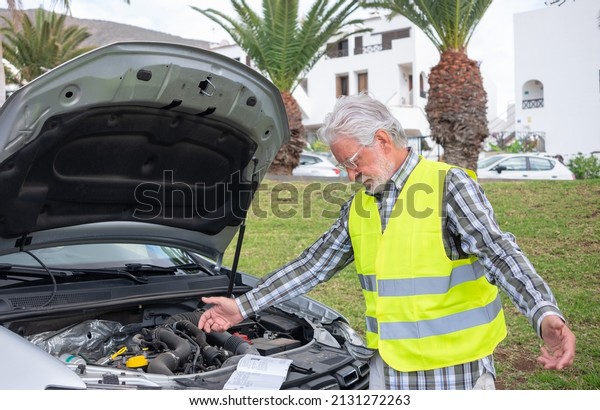 Senior driver stopped in the street for a car\
breakdown with the hood open wearing the yellow vest waiting for\
roadside assistance. Look at the instruction booklet to understand\
what is broken