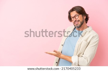 Senior dutch man isolated on pink background presenting an idea while looking smiling towards