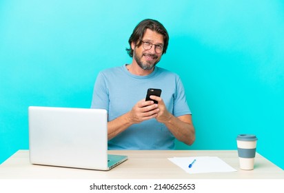 Senior dutch man in a table with a laptop isolated on blue background sending a message with the mobile