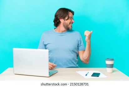 Senior dutch man in a table with a laptop isolated on blue background pointing to the side to present a product
