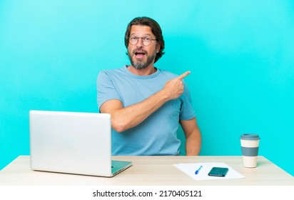 Senior dutch man in a table with a laptop isolated on blue background surprised and pointing side
