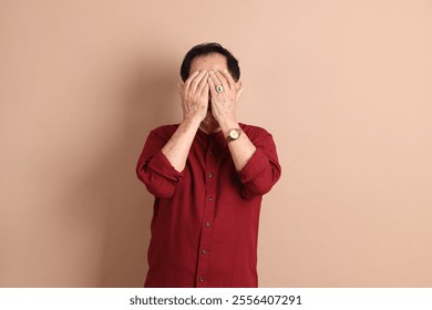 Senior East Asian man with a deep red shirt standing against a brown background. - Powered by Shutterstock