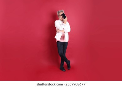 A senior East Asian man in a white shirt stands against a deep red background. - Powered by Shutterstock