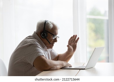 Senior Elder 80s Lonely Man In Headphones With Microphone Waving Hand At Laptop Screen, Making Video Call, Saying Hello, Talking, To Family, Sitting At Table With Computer. Side View