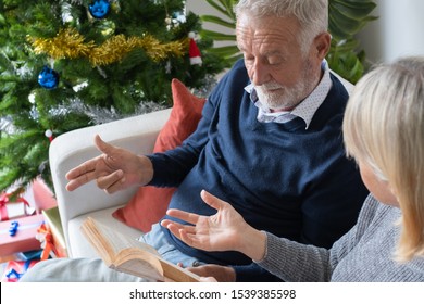 Senior Elderly Caucasian Old Man And Woman Reading Book And Sitting On Sofa To Discussion Together In Living Room That Decorated For Christmas Festival Day In The Morning, Retirement Family Concep