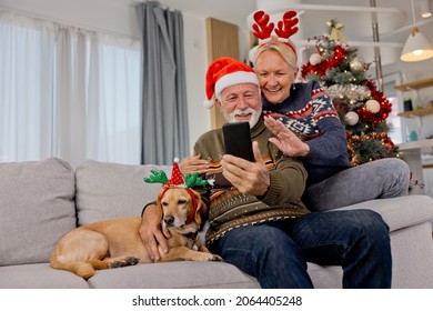 Senior elderly couple having video call over mobile phone sitting on sofa at home with their yellow dog and wearing Christmas sweaters during festive season - Powered by Shutterstock