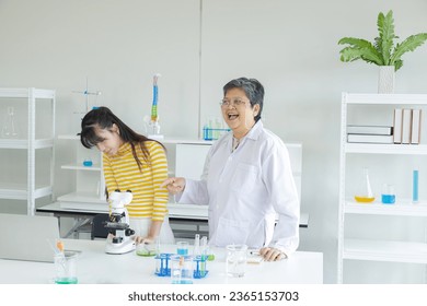 Senior elderly employment, happy teacher wearing lab coat teaching in science laboratory with cheerful Asian students, pupil girl volunteer standing in front of classroom using microscope experiment - Powered by Shutterstock