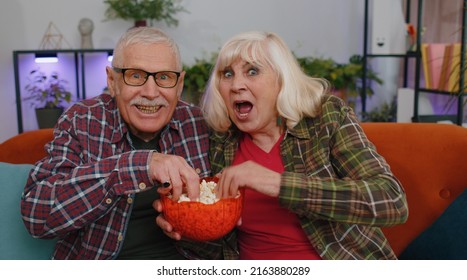 Senior Elderly Family Couple Grandparents Man Woman Watching Interesting Television Movie, Football, Sport Games, Eating Popcorn. Mature Grandmother Grandfather Enjoying Leisure Time On Sofa At Home