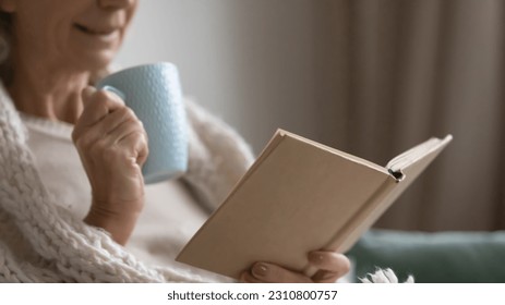 Senior elderly lady wrapped in woolen comforter reading learning book, getting knowledge over cup of hot drink. Mature woman enjoying leisure on couch, morning coffee, studying at home. Close up - Powered by Shutterstock