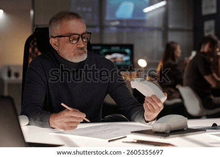 Senior engineer in eyeglasses and pullover sitting by workplace in bureau and looking at car model while sketching it on paper