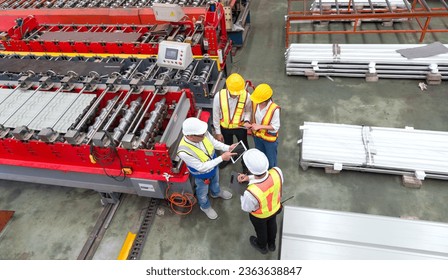 Senior engineer manager trains new employees within the metal sheet factory. Everyone wear safety vest and hardhat. Large machines and metal sheets are in the working area.