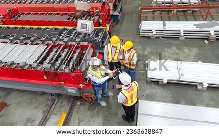 Senior engineer manager trains new employees within the metal sheet factory. Everyone wear safety vest and hardhat. Large machines and metal sheets are in the working area.