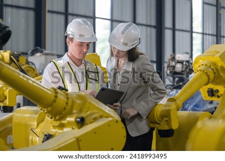 Senior engineer provides guidance to a trainee amidst robotic arms in a high-tech warehouse factory setting.