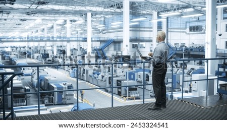 Senior Engineer Standing on a Platform, Using Laptop Computer at an Electronics Factory. Machines are Undergoing Maintenance, Specialist Monitoring the Progress Through Online Software