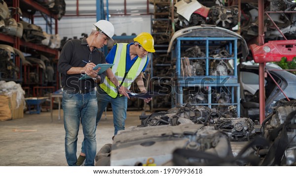 senior engineer using laptop computer checking\
list old used car engine with young asian worker in machinery parts\
shop warehouse. old manager find vehicle automobile machine in\
Recycle motor factory
