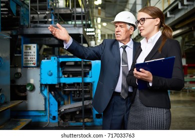 Senior entrepreneur wearing hardhat and his attractive young assistant manager carrying out inspection at production department of modern plant