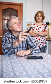 Senior European Couple Relationships, Man Watching Tv With Remote Control While His Angry Wife Standing Behind In Kitchen