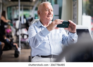 Senior European man sitting on seat inside tram and taking photographs with his smartphone.