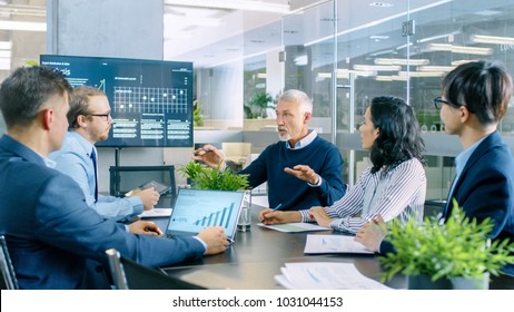 Senior Executive Explains Company's Vision And Potential To His Employees. They Are Sitting At Big Table In Meeting Room. TV Screen On The Wall Shows Corporate Growth.