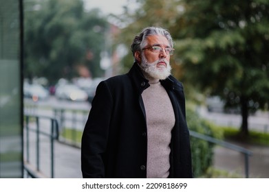 Senior Executive Walking Outside And Going To Work At The Business Center. Happy Elderly Businessman Standing In Front Of The Office Building And Looking Away While Wearing Black Coat. Copy Space.