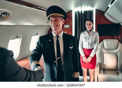 Senior Experienced Pilot In Uniform And Beautiful Female Flight Attendant In Private Jet. Standing In Aisle Together And Meeting Passengers