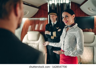 Senior Experienced Pilot In Uniform And Beautiful Female Flight Attendant In Private Jet. Standing In Aisle Together And Meeting Passengers