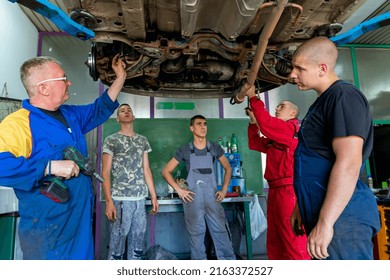 Senior Experienced Teacher Standing Underneath Car On Hydraulic Ramp And Teaching Automotive Trade To His Students In The Vocational School