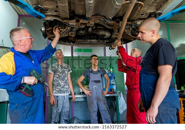 Senior experienced teacher standing underneath car\
on hydraulic ramp and teaching automotive trade to his students in\
the vocational school