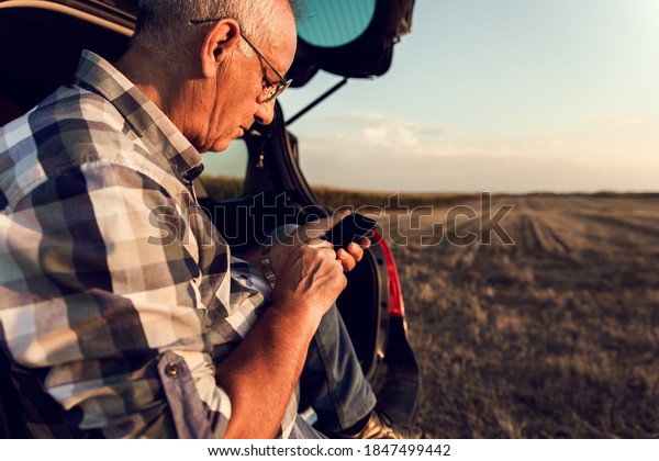 Senior farmer siting in car boot\
in the field, looking at phone after wheat harvest at\
sunset.
