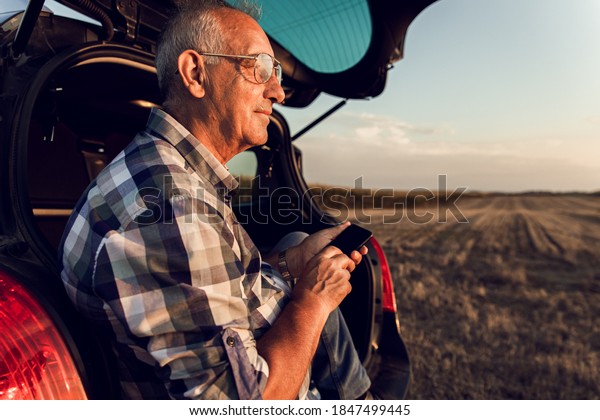Senior farmer siting in car boot, watching wheat\
field after harvest at\
sunset.