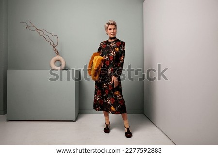 Senior fashion female model standing full length in studio wears black maxi floral dress and socks with shoes. Mature woman with short hair
