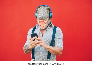 Senior Fashion Man Wearing T-rex Mask Using Mobile Smartphone Listening Music With Headphones - Crazy Bizarre Hipster Guy Having Fun With New Technology -  Funny And Absurd Surreal Concepts