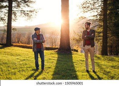 Senior father and his son standing by tree at sunset, looking at each other.