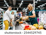 Senior female cashier scanning groceries of a customer at checkout counter in a supermarket, with a senior man pushing his shopping cart and waiting to pay for his groceries