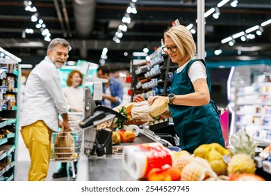 Senior female cashier scanning groceries of a customer at checkout counter in a supermarket, with a senior man pushing his shopping cart and waiting to pay for his groceries - Powered by Shutterstock